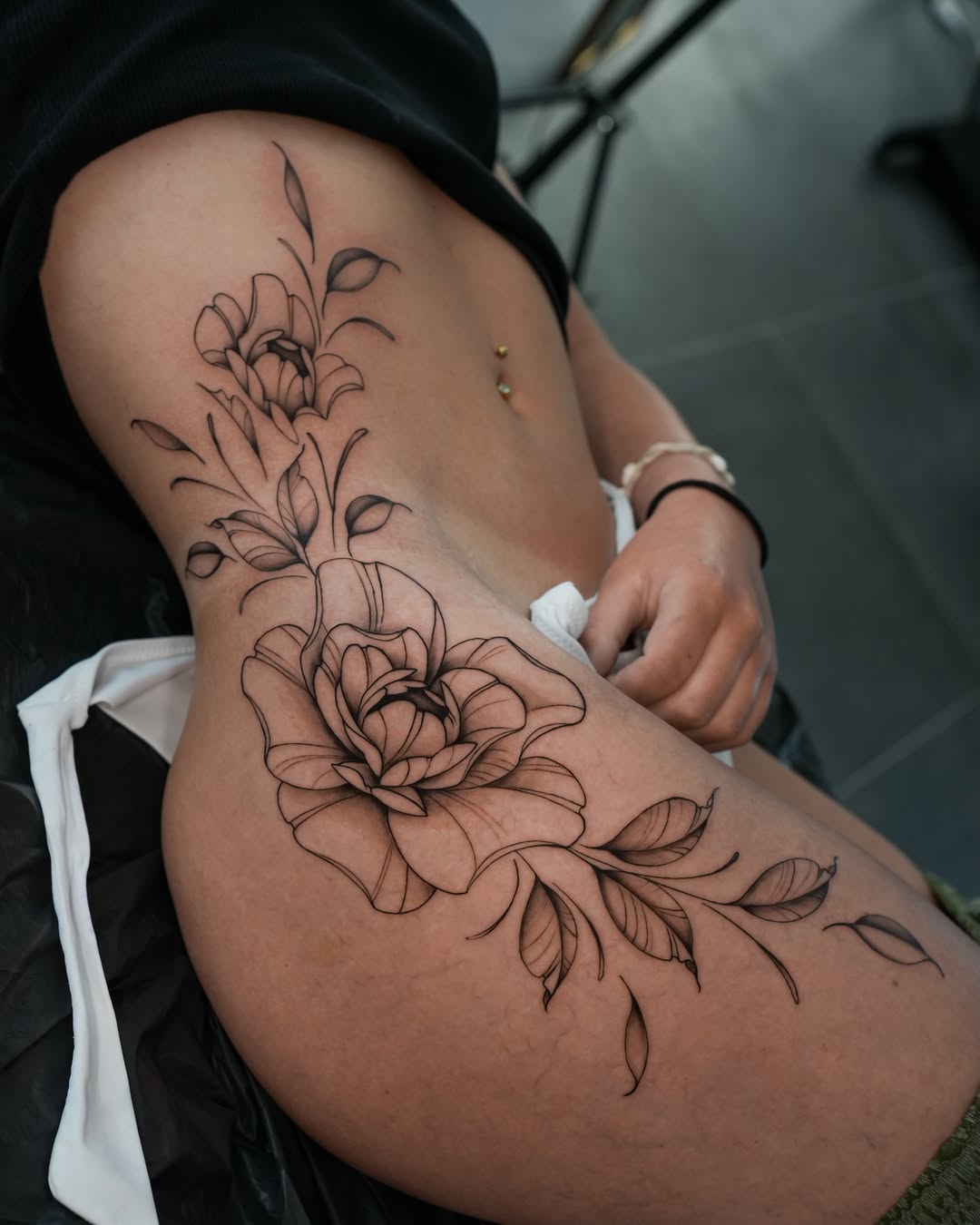 Filigranes Fineline Blumen-Tattoo an der Hüfte mit Bauchnabelpiercing